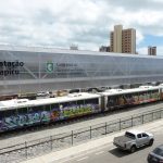 Veículo leve sobre trilhos é visto de lado, parado na estação Papicu, em Fortaleza. O céu, ao fundo, tem muitas nuvens, embora o dia esteja ensolarado. O veículo é branco com alguns grafites ao longo de sua extensão. Mais abaixo na imagem, pode-se ver a rua paralela a estação com veículos circulando.