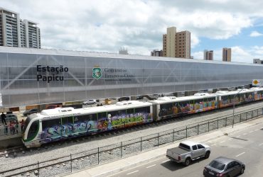Veículo leve sobre trilhos é visto de lado, parado na estação Papicu, em Fortaleza. O céu, ao fundo, tem muitas nuvens, embora o dia esteja ensolarado. O veículo é branco com alguns grafites ao longo de sua extensão. Mais abaixo na imagem, pode-se ver a rua paralela a estação com veículos circulando.
