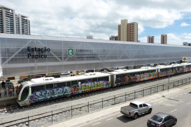 Veículo leve sobre trilhos é visto de lado, parado na estação Papicu, em Fortaleza. O céu, ao fundo, tem muitas nuvens, embora o dia esteja ensolarado. O veículo é branco com alguns grafites ao longo de sua extensão. Mais abaixo na imagem, pode-se ver a rua paralela a estação com veículos circulando.