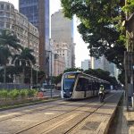 VLT carioca visto no entardecer do Rio de Janeiro, circulando na Cinelândia. Ao fundo, do lado esquerdo na diagonal, se vê os prédios da região. Do lado direito, as árvoes, Ao lado do trem, do lado direito está um motoqueiro circulando com o veículo.