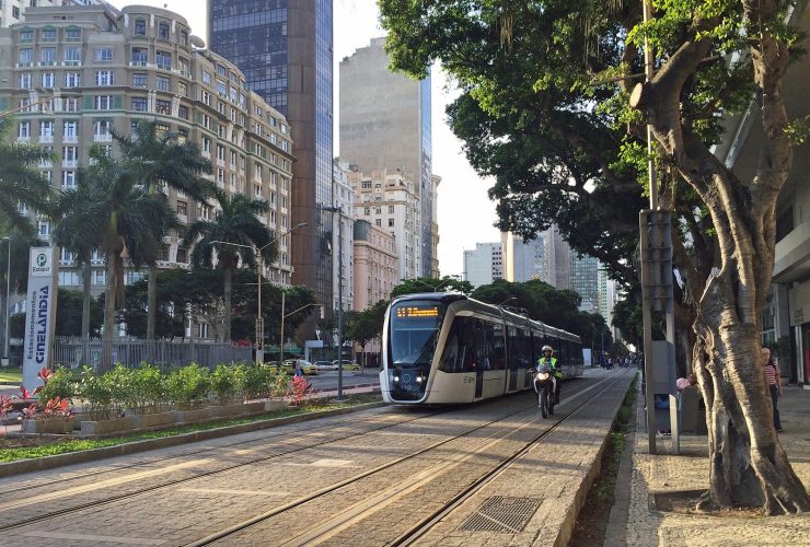 VLT carioca visto no entardecer do Rio de Janeiro, circulando na Cinelândia. Ao fundo, do lado esquerdo na diagonal, se vê os prédios da região. Do lado direito, as árvoes, Ao lado do trem, do lado direito está um motoqueiro circulando com o veículo.