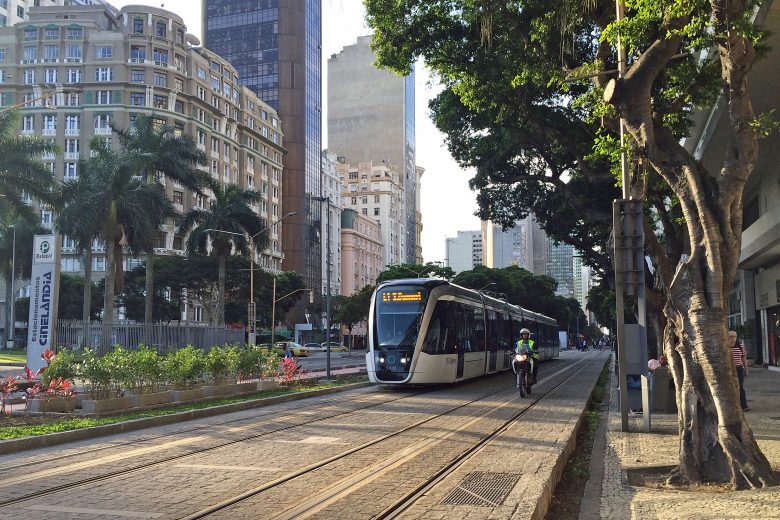 VLT carioca visto no entardecer do Rio de Janeiro, circulando na Cinelândia. Ao fundo, do lado esquerdo na diagonal, se vê os prédios da região. Do lado direito, as árvoes, Ao lado do trem, do lado direito está um motoqueiro circulando com o veículo.