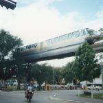 Monotrilho de Poços de Caldas operando sobre uma avenida da cidade. O veículo é cinza com uma faixa amarela na lateral, com detalhes em azul nas extremidades. Ao lado da viga que mantem o monotrilho no ar, pairam árvores. Abaixo, na rua, trafega uma moto com o motorista sem capacete.