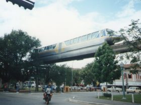 Monotrilho de Poços de Caldas operando sobre uma avenida da cidade. O veículo é cinza com uma faixa amarela na lateral, com detalhes em azul nas extremidades. Ao lado da viga que mantem o monotrilho no ar, pairam árvores. Abaixo, na rua, trafega uma moto com o motorista sem capacete.