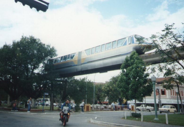 Monotrilho de Poços de Caldas operando sobre uma avenida da cidade. O veículo é cinza com uma faixa amarela na lateral, com detalhes em azul nas extremidades. Ao lado da viga que mantem o monotrilho no ar, pairam árvores. Abaixo, na rua, trafega uma moto com o motorista sem capacete.