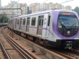 Frente do trem é vista de frente, em movimento circulando na linha lilas. O trem é cinza metálico e lilás. Foto diurna.