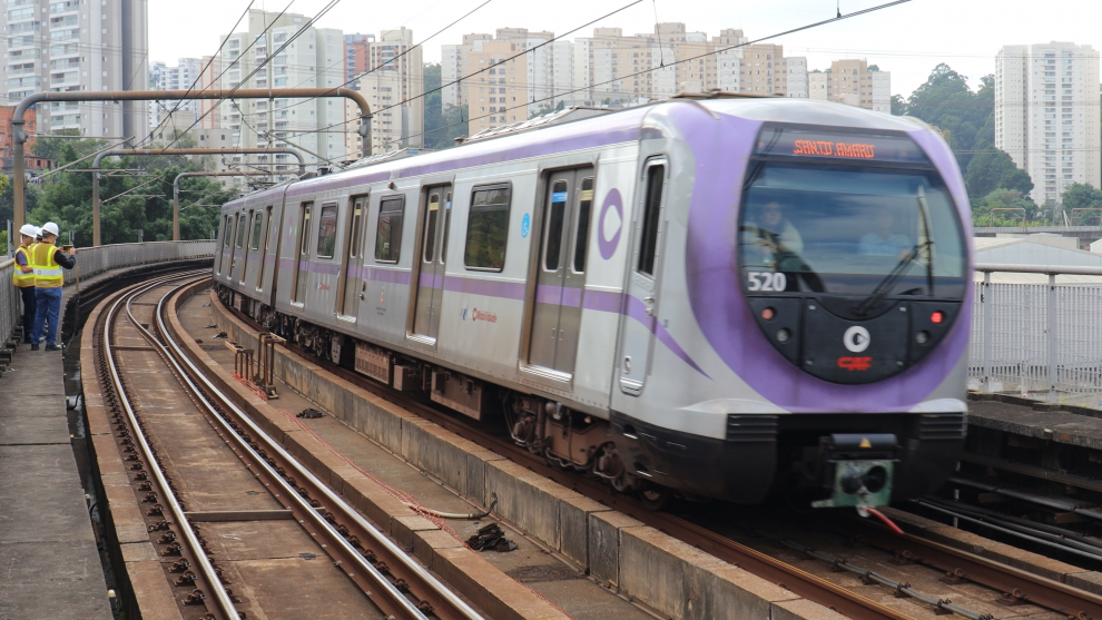 Frente do trem é vista de frente, em movimento circulando na linha lilas. O trem é cinza metálico e lilás. Foto diurna.