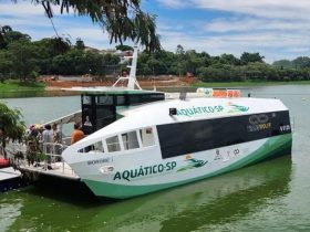 Barco "Bororé I" durante demonstração na Represa Billings. O barco está visto na diagonal dianteira sobre a água esverdeada. Ao lado há uma plataforma de embarque flutuante. Há algumas pessoas na parte dianteira do barco. Ao fundo pode ser vista uma margem.