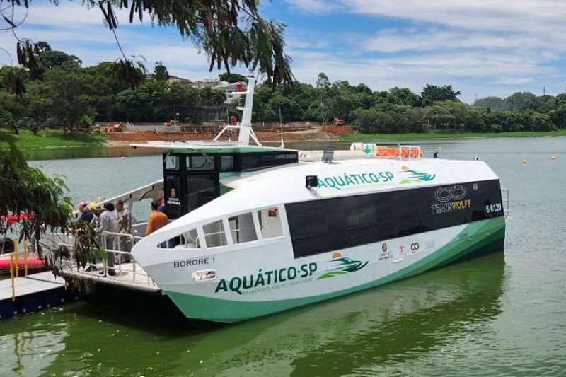 Barco "Bororé I" durante demonstração na Represa Billings. O barco está visto na diagonal dianteira sobre a água esverdeada. Ao lado há uma plataforma de embarque flutuante. Há algumas pessoas na parte dianteira do barco. Ao fundo pode ser vista uma margem.