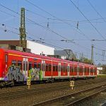 Trem vermelho com grafites no primeiro carro, circula por trilhos abaixo de um sistema de catenária. O trem é visto pela frente, na diagonal com o céu azul claro ao fundo. O chão é com pedras de cor marrom.