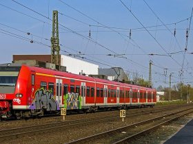 Trem vermelho com grafites no primeiro carro, circula por trilhos abaixo de um sistema de catenária. O trem é visto pela frente, na diagonal com o céu azul claro ao fundo. O chão é com pedras de cor marrom.