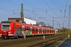 Trem vermelho com grafites no primeiro carro, circula por trilhos abaixo de um sistema de catenária. O trem é visto pela frente, na diagonal com o céu azul claro ao fundo. O chão é com pedras de cor marrom.
