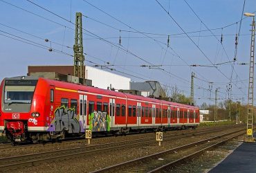 Trem vermelho com grafites no primeiro carro, circula por trilhos abaixo de um sistema de catenária. O trem é visto pela frente, na diagonal com o céu azul claro ao fundo. O chão é com pedras de cor marrom.