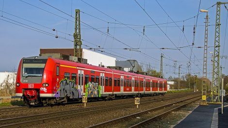 Trem vermelho com grafites no primeiro carro, circula por trilhos abaixo de um sistema de catenária. O trem é visto pela frente, na diagonal com o céu azul claro ao fundo. O chão é com pedras de cor marrom.