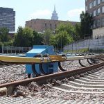 Terceiro trilho (com cobertura amarela), ao lado esquerdo dos trilhos, no sistema ferroviário próximo a estação Sokol, em Moscou, Rússia. Ao fundo, vê-se um céu azul claro e edifícios da cidade.