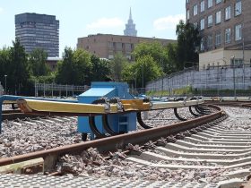 Terceiro trilho (com cobertura amarela), ao lado esquerdo dos trilhos, no sistema ferroviário próximo a estação Sokol, em Moscou, Rússia. Ao fundo, vê-se um céu azul claro e edifícios da cidade.