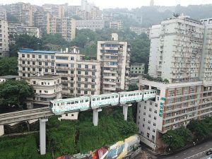 Monotrilho de Chongqing na China em operação. Na imagem o monotrilho de cores claras circula suspenso enquanto está entrando dentro de um edifício residencial. A vista á lateral. Ao fundo, estão outros edifícios residenciais em uma encosta cercada de vegetação verde. A imagem é diurna com neblina.