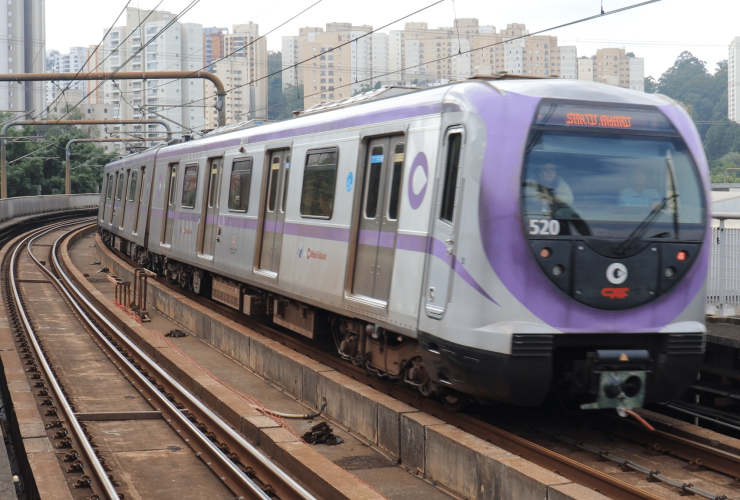 Frente do trem é vista de frente, em movimento circulando na linha lilas. O trem é cinza metálico e lilás. Foto diurna.