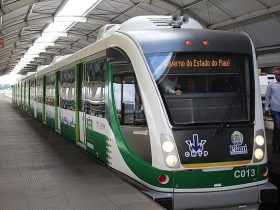 VLT em operação no Metrô de Teresina durante o dia, parado em uma estação com as portas fechadas. Na imagem, o trem é visto na diagonal, de frente. O veículo é branco com detalhes em verde. A estação tem teto em formato circular com uma abertura no alto que permite entrada de luz natural. Há uma pessoa de pé, do lado direito da imagem vestindo camisa clara e calça jeans. No letreiro do trem, pode-se ler "Governo do Estado do Piauí".