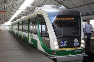 VLT em operação no Metrô de Teresina durante o dia, parado em uma estação com as portas fechadas. Na imagem, o trem é visto na diagonal, de frente. O veículo é branco com detalhes em verde. A estação tem teto em formato circular com uma abertura no alto que permite entrada de luz natural. Há uma pessoa de pé, do lado direito da imagem vestindo camisa clara e calça jeans. No letreiro do trem, pode-se ler "Governo do Estado do Piauí".